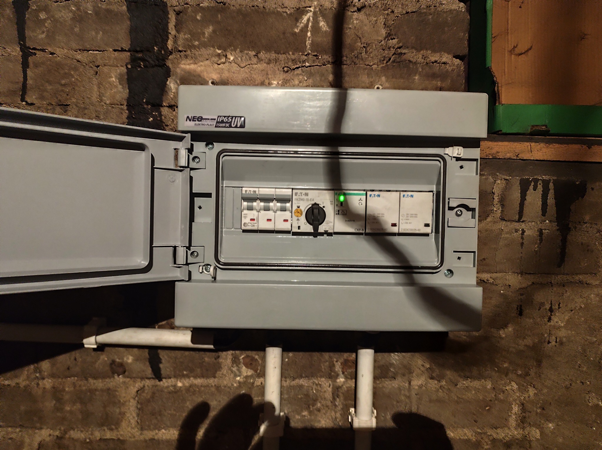 Szara skrzynka elektryczna z otwartymi drzwiczkami, zamontowana na ceglanej ścianie. Widoczne bezpieczniki i przełączniki wewnątrz. Oświetlenie punktowe.