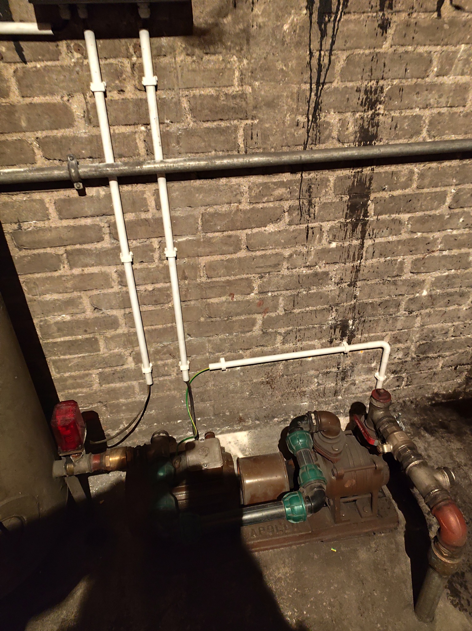 Instalacja wodna z pompą Apollo na tle ceglanej ściany z zaciekami. Widoczne białe rury PCV i metalowe elementy. Niskie oświetlenie podkreśla surowy charakter piwnicy.