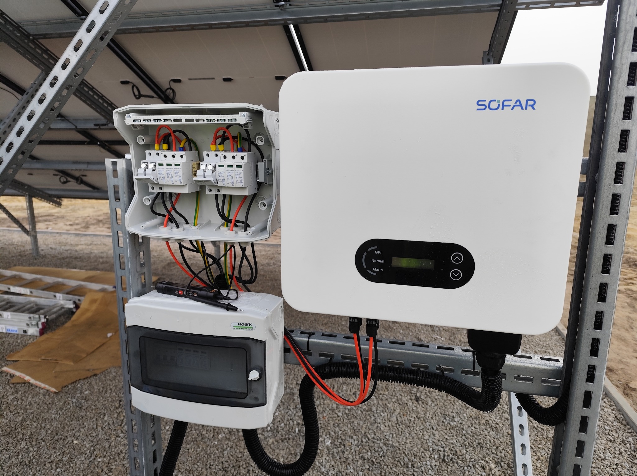 Biały inwerter SOFAR z podłączonymi przewodami i skrzynką bezpiecznikową na metalowej konstrukcji, instalacja fotowoltaiczna w trakcie montażu, widoczne elementy pomiarowe.