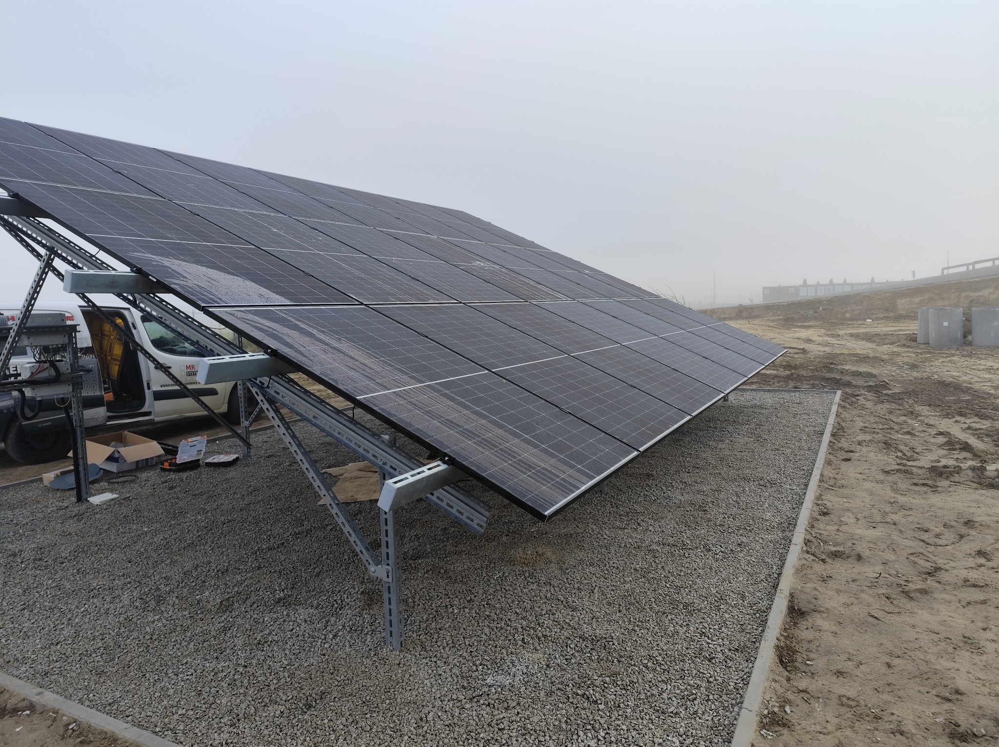 Instalacja paneli fotowoltaicznych na gruncie w Otwocku. Widoczny częściowo samochód dostawczy i narzędzia. Mglista pogoda, teren przygotowany pod inwestycję.