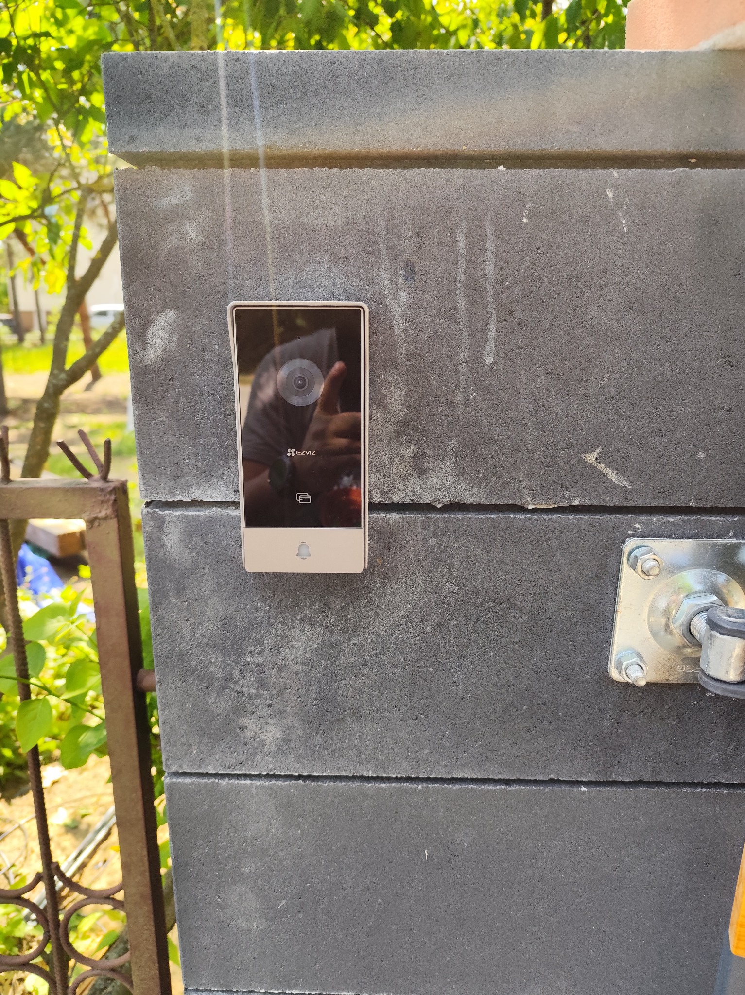 Zamontowany wideodomofon marki Ezviz na szarym, betonowym słupku ogrodzenia. Widoczny fragment furtki i zieleń w tle. Słoneczny dzień.
