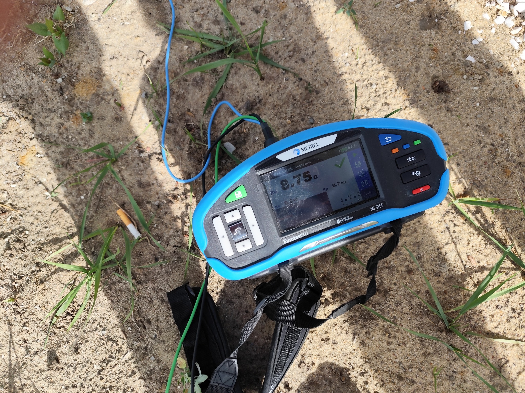 Miernik rezystancji uziemienia Metrel MI 3155 na piasku z trawą, wskazujący wynik 8.75 Ω. Widoczne przewody pomiarowe i niedopałek papierosa obok.