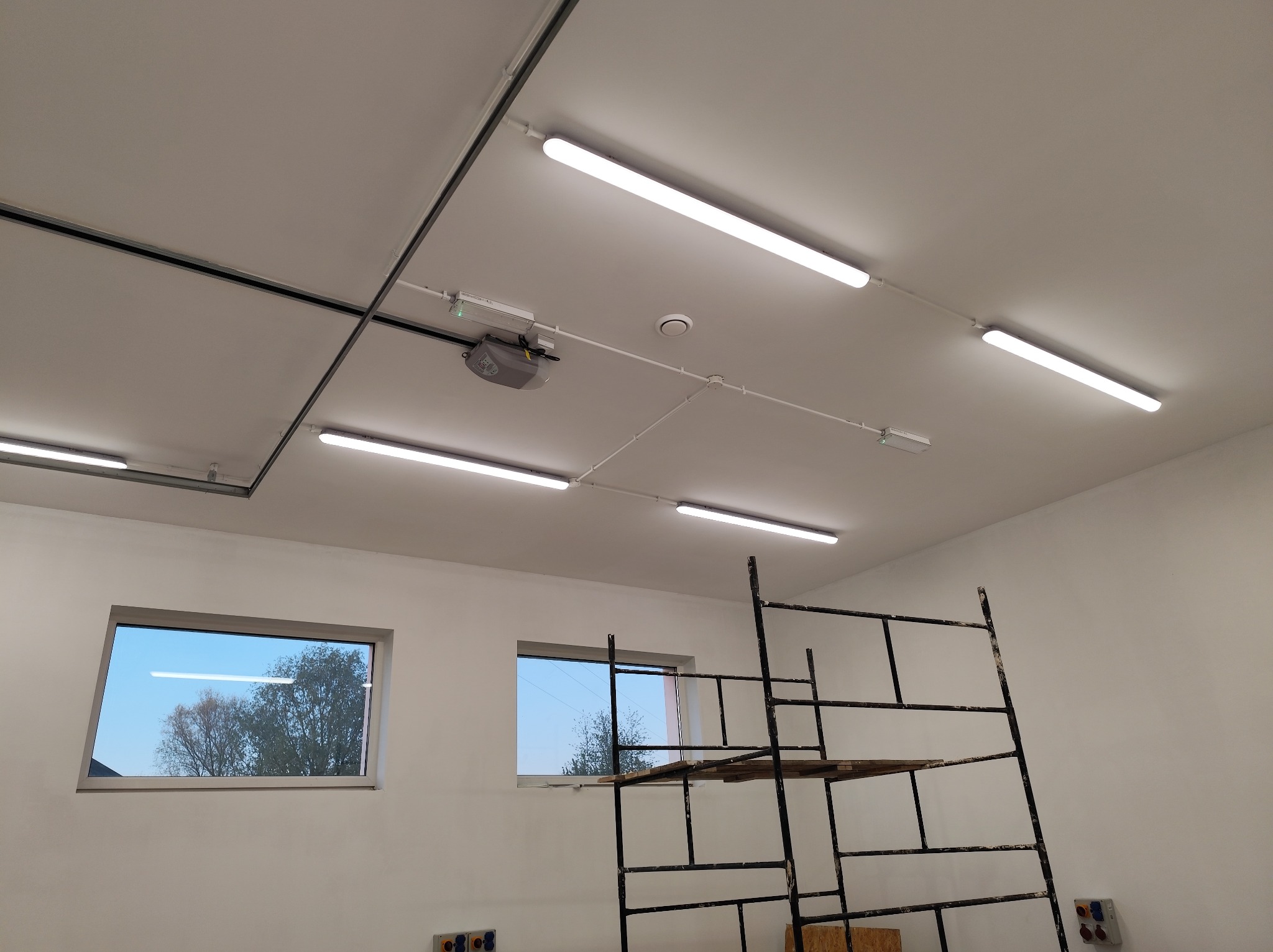 Białe, podłużne lampy LED zamontowane na suficie w nowym pomieszczeniu. Widoczne okna i rusztowanie malarskie. Instalacja oświetlenia w trakcie prac wykończeniowych.