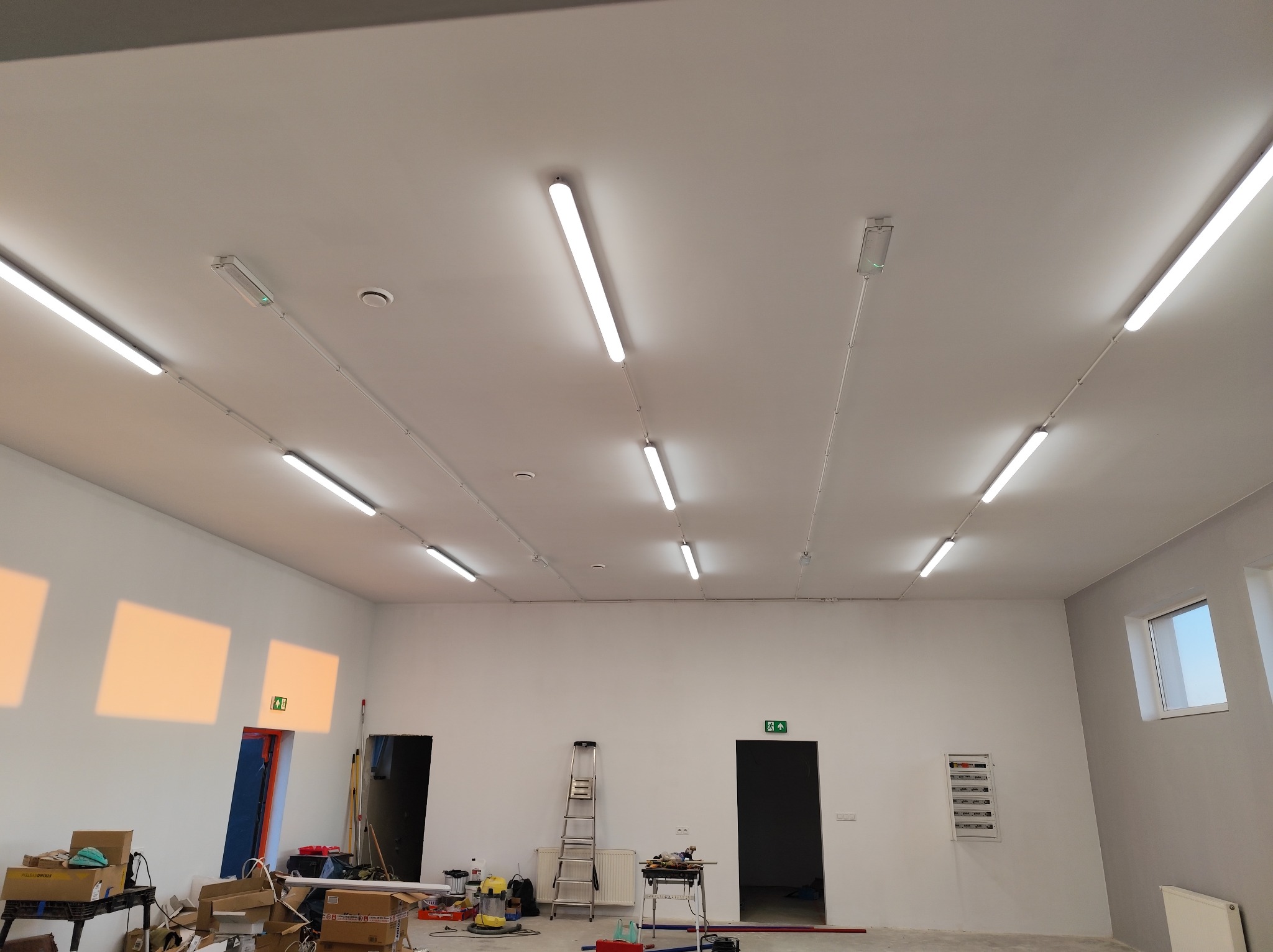 Białe świetlówki LED zamontowane na suficie w nowym, przestronnym pomieszczeniu z białymi ścianami. Widoczne skrzynki elektryczne, drabina i narzędzia.