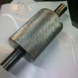 Metalowy cylinder z radełkowaną powierzchnią i dwoma przymocowanymi wałkami, leżący na białym arkuszu. Detal wykonany z precyzją.