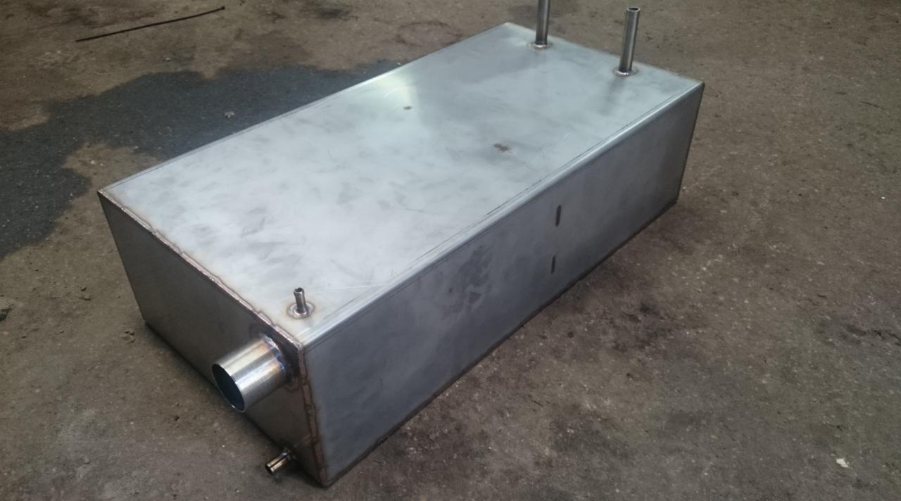 Prostokątny, metalowy zbiornik z rurkami, wykonany ze stali nierdzewnej, leżący na betonowej podłodze. Widoczne spawy i detale wykończenia.