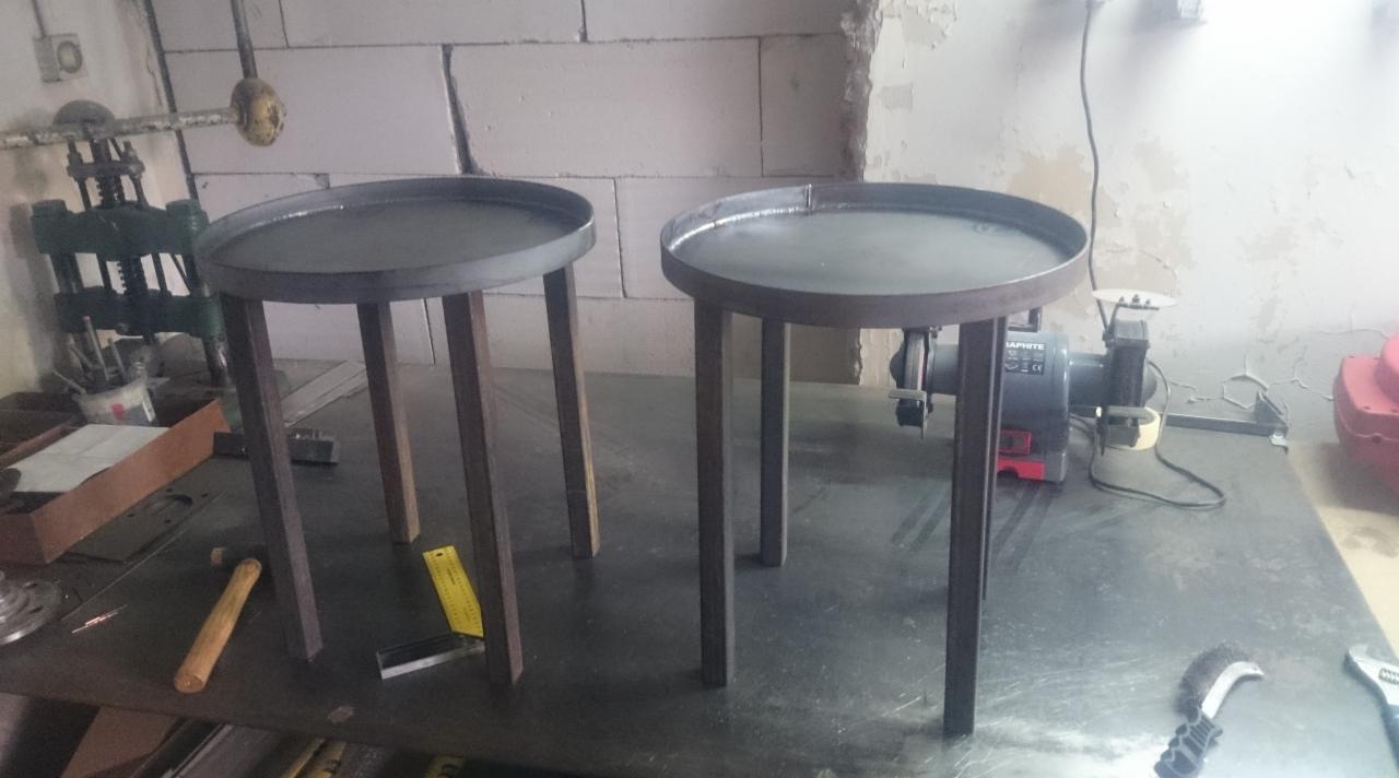 Dwa metalowe stoliki z okrągłymi blatami na warsztatowym stole, obok narzędzia i szlifierka. Blat stołu z widocznymi śladami obróbki.
