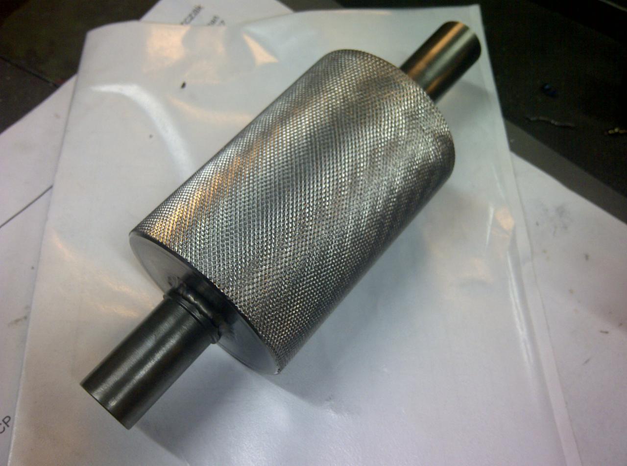 Metalowy cylinder z radełkowaną powierzchnią i dwoma przymocowanymi wałkami, leżący na białym arkuszu. Detal wykonany z precyzją.