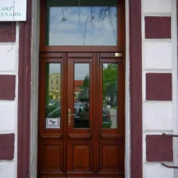 Brązowe drzwi wejściowe z przeszkleniami, otoczone budynkiem z biało-bordową elewacją. Nad drzwiami dodatkowe okno doświetlające.