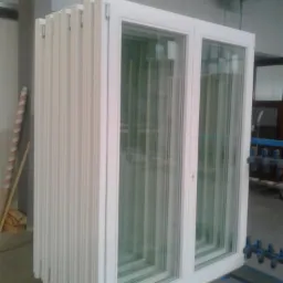 Białe, podwójne okno z szybami w magazynie, ustawione obok rzędu identycznych ram okiennych, gotowych do montażu. Widoczne zawiasy i detale wykończenia.