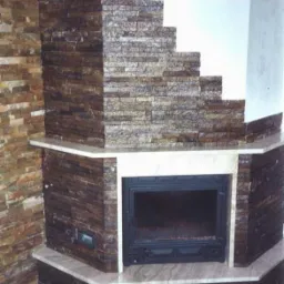 Kamienny kominek narożny z jasnym blatem, wykończony w odcieniach brązu i beżu, z widocznym wkładem kominkowym i kratkami wentylacyjnymi.