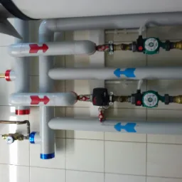 Szara instalacja CO z rur, zaworów i pomp obiegowych, strzałki wskazują kierunek przepływu. Widoczne manometry i odpowietrzniki. Tło stanowią białe płytki.