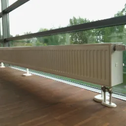 Długi, biały grzejnik panelowy na niskich nóżkach, zamontowany pod oknem w nowoczesnym wnętrzu z drewnianą podłogą. Widok na zieleń za oknem.