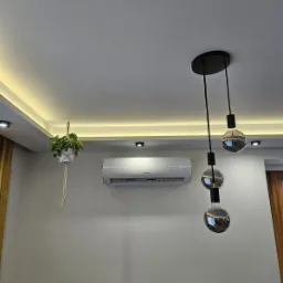 Nowoczesny salon z zamontowanym klimatyzatorem marki Gree, wiszącą rośliną w makramie i designerską lampą z trzema żarówkami w stylu retro.