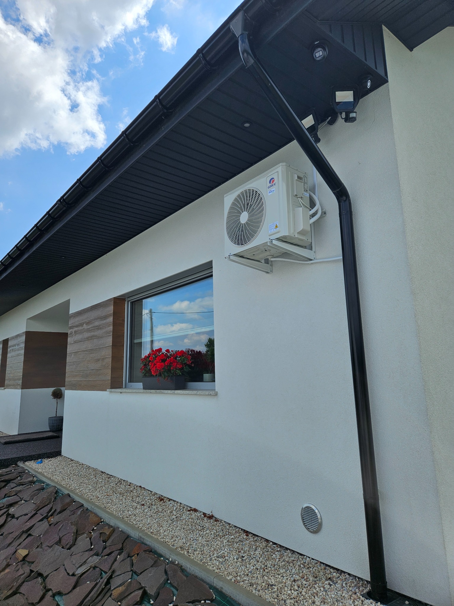 Zewnętrzna jednostka klimatyzacji zamontowana na białej ścianie domu z widocznymi elementami instalacji, czarną rynną i kwiatami w oknie.