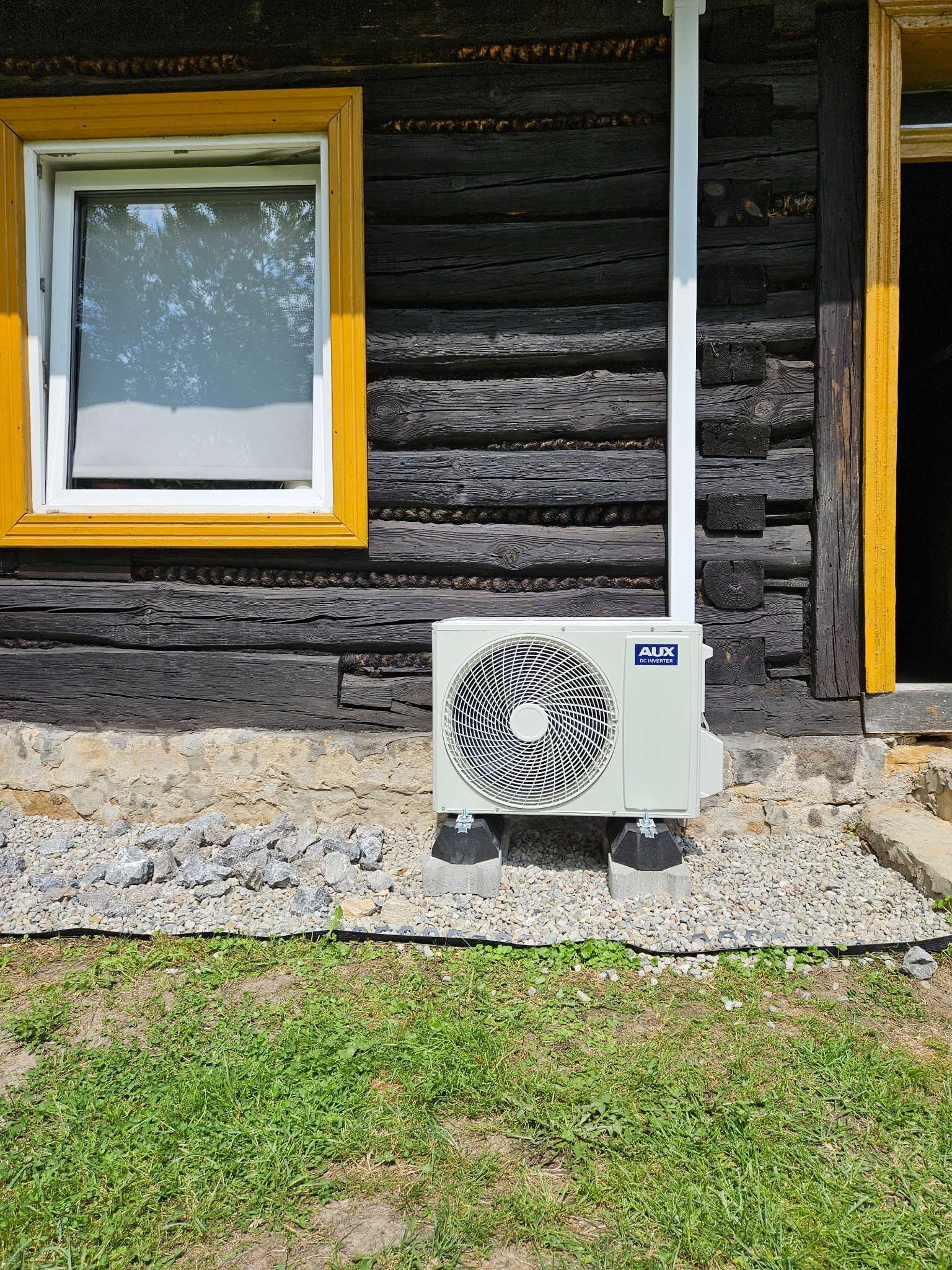Zewnętrzna jednostka klimatyzacji zamontowana na betonowych podstawach przy drewnianym domu z żółtymi ramami okiennymi i kamiennym fundamentem.