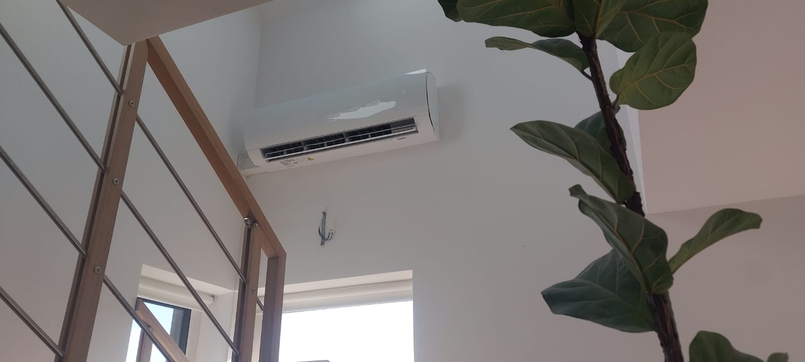 Biały klimatyzator ścienny w minimalistycznym wnętrzu z drewnianą balustradą i rośliną doniczkową, widoczne ślady po folii ochronnej.