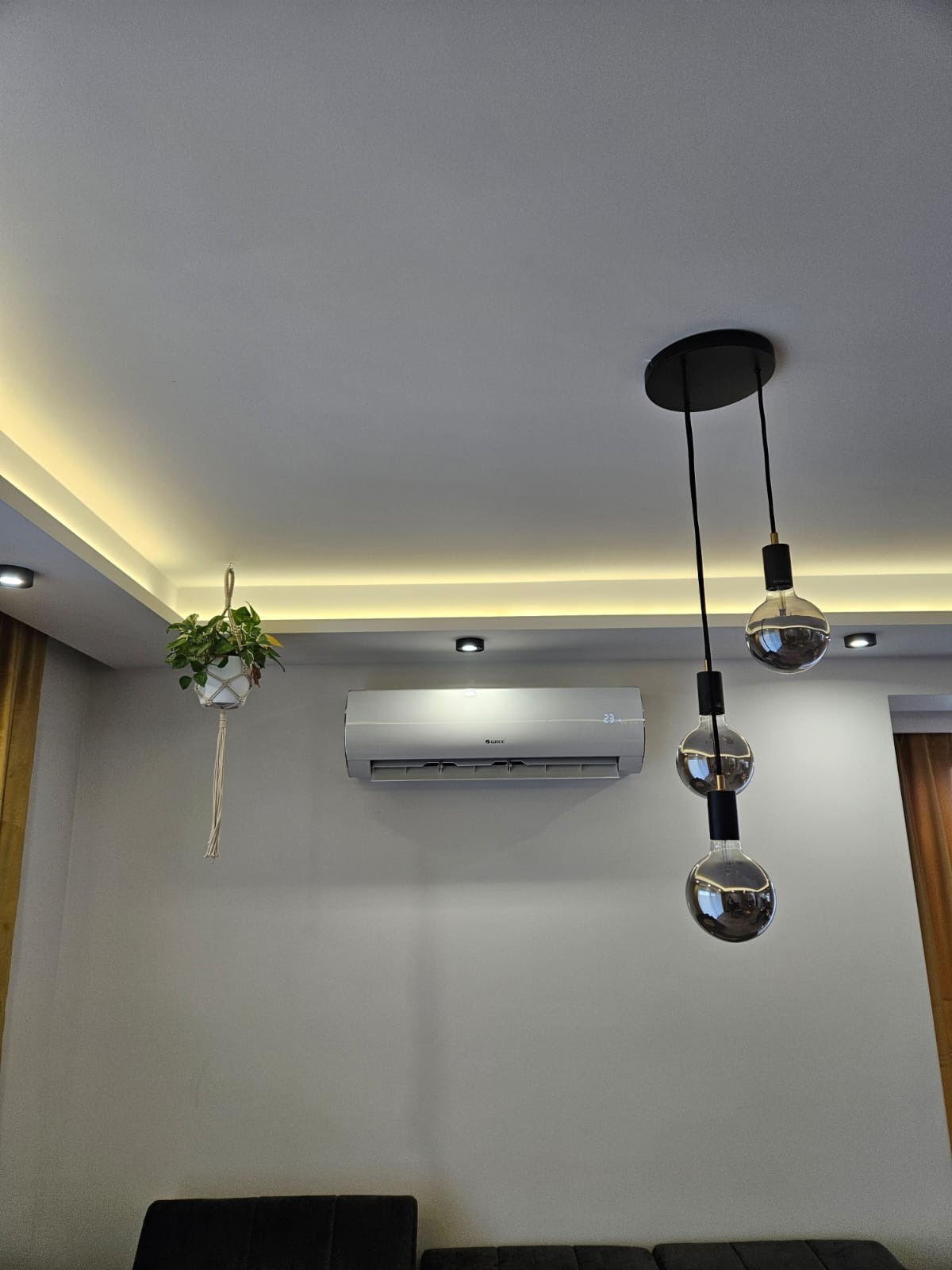 Nowoczesny salon z zamontowanym klimatyzatorem marki Gree, wiszącą rośliną w makramie i designerską lampą z trzema żarówkami w stylu retro.