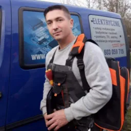 Elektryk w stroju roboczym, z torbą na narzędzia, na tle niebieskiego samochodu z reklamą usług. Widoczny numer telefonu i zakres usług.