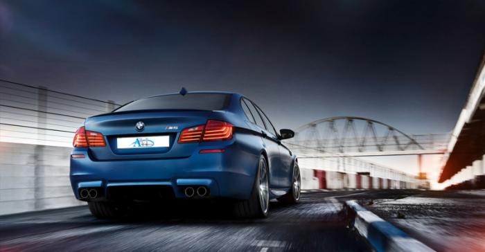 Błękitny samochód marki BMW, model M5, widziany z tyłu, na torze wyścigowym z mostem w tle, w dynamicznym ujęciu z efektem rozmycia ruchu.