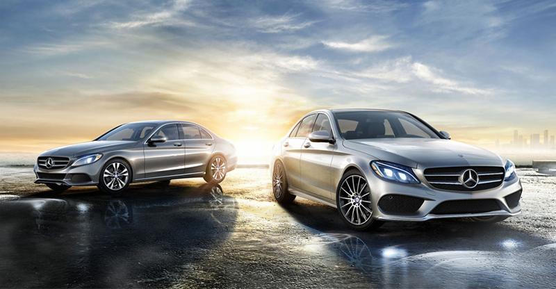 Dwa srebrne samochody Mercedes klasy C na mokrej nawierzchni z odbiciem, w tle zachód słońca i panorama miasta.