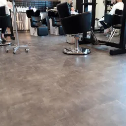 Szara posadzka w salonie fryzjerskim z widocznymi krzesłami i stanowiskami do mycia włosów, ułożona z płytek imitujących beton.