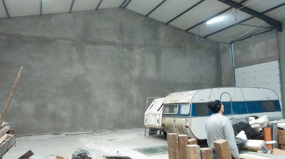 Świeżo otynkowana, gładka ściana w hali przemysłowej, z widocznymi elementami konstrukcji dachu i zaparkowaną przyczepą kempingową, obok której stoi osoba w czapce.