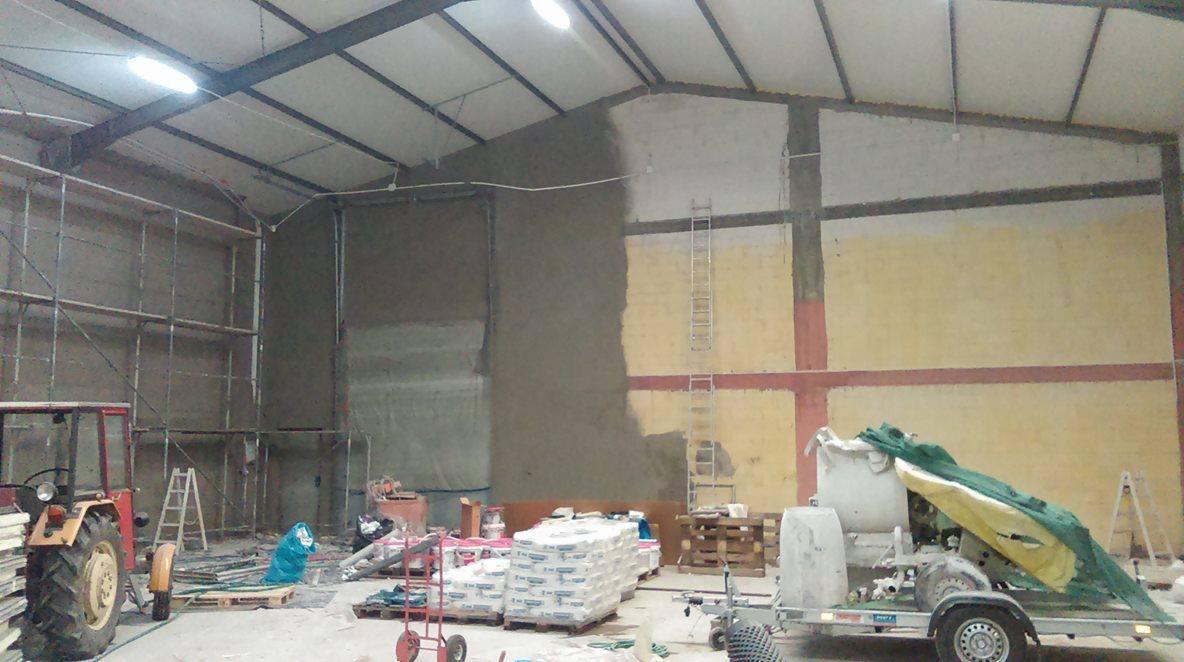 Wnętrze hali w trakcie prac tynkarskich: częściowo otynkowana ściana, rusztowania, maszyna tynkarska na przyczepie, worki z zaprawą i traktor z przodu po lewej.
