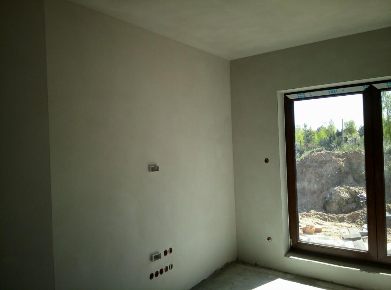 Świeżo położone, jasnoszare tynki maszynowe na ścianach w pokoju, widoczne puszki elektryczne i okno z widokiem na plac budowy.