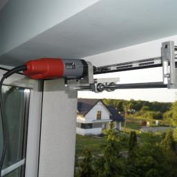 Nawiewniki Białystok - Montaż automatycznego otwierania okna balkonowego za pomocą wiertarki z adapterem. Widok na zewnątrz, dom w tle. Specyficzny adapter do wiertarki.