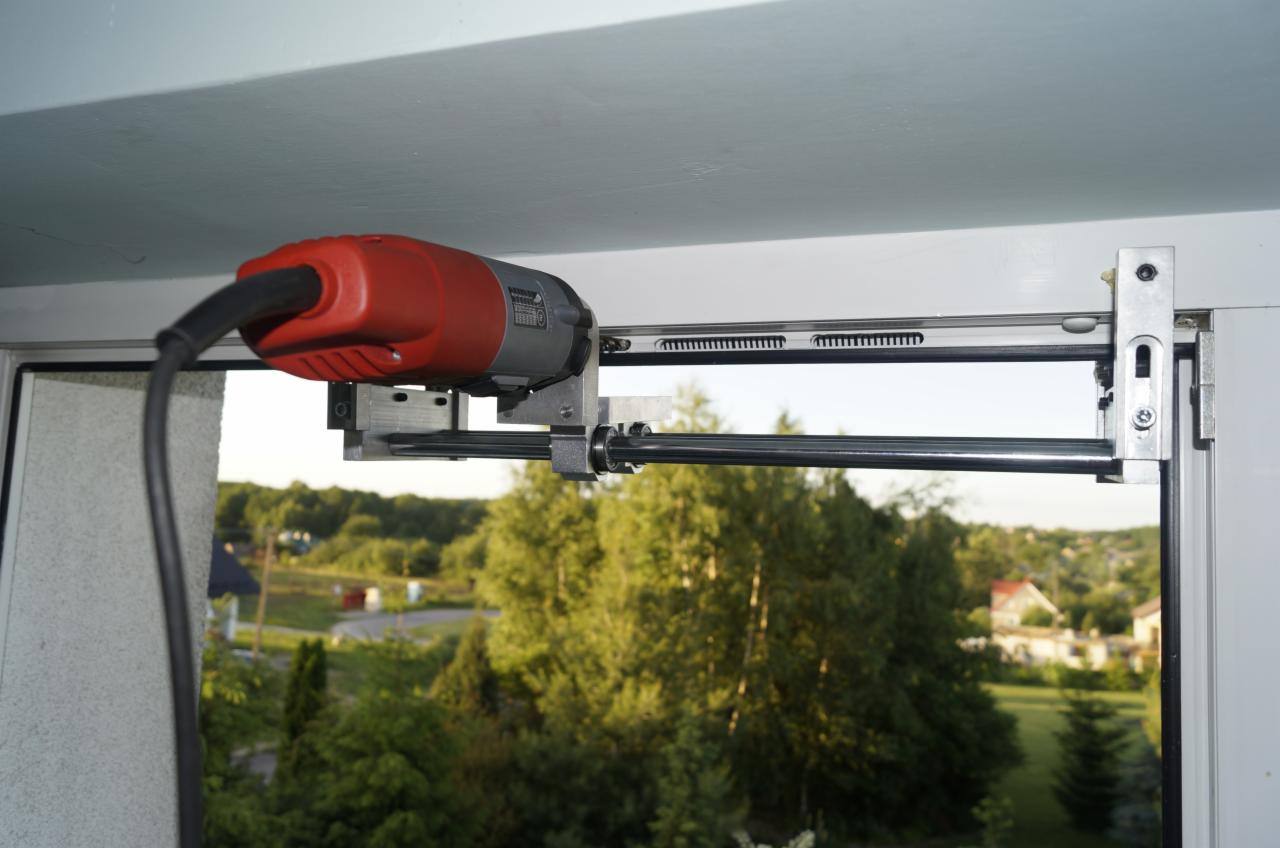 Specjalistyczne narzędzie do naprawy okien zamontowane na ramie okiennej, widok na zewnątrz przez okno. Precyzyjny mechanizm w trakcie pracy.