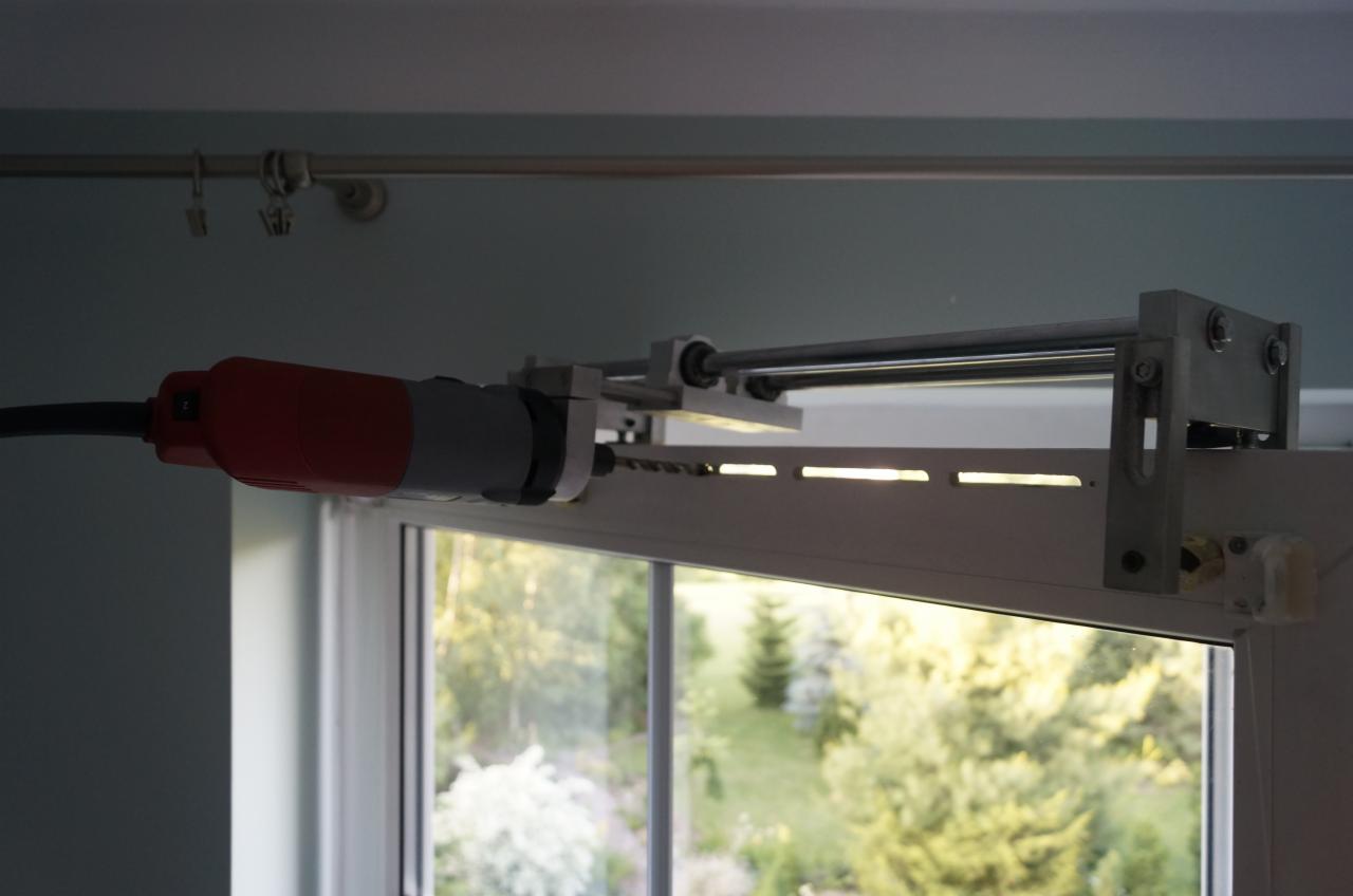 Wiertarka z wiertłem montowana do ramy okiennej z roletami zewnętrznymi, widok z bliska na mechanizm. Rolety częściowo opuszczone, widok na zieleń za oknem.