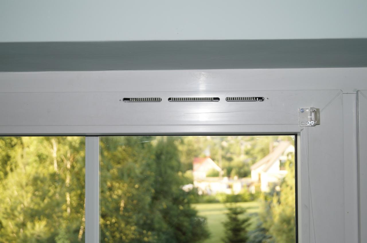 Białe okno PCV z widocznymi nawiewnikami szczelinowymi w górnej ramie, widok na zewnątrz z zieleniejącym ogrodem i domami w oddali.