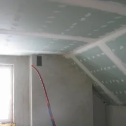 Poddasze z oknem: płyty gipsowo-kartonowe na skosach, szpachlowane łączenia, widoczne rury instalacyjne przy ścianie. Etap wykończenia wnętrza.