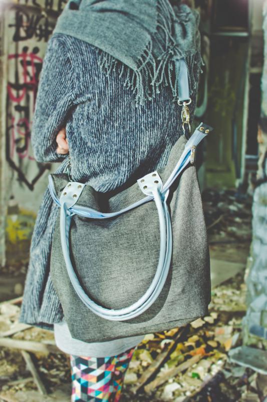 Szara torba na ramię z jasnoniebieskimi uchwytami, noszona przez osobę w szarym płaszczu na tle ściany z graffiti. Detal torby, widoczne uchwyty i zamek.