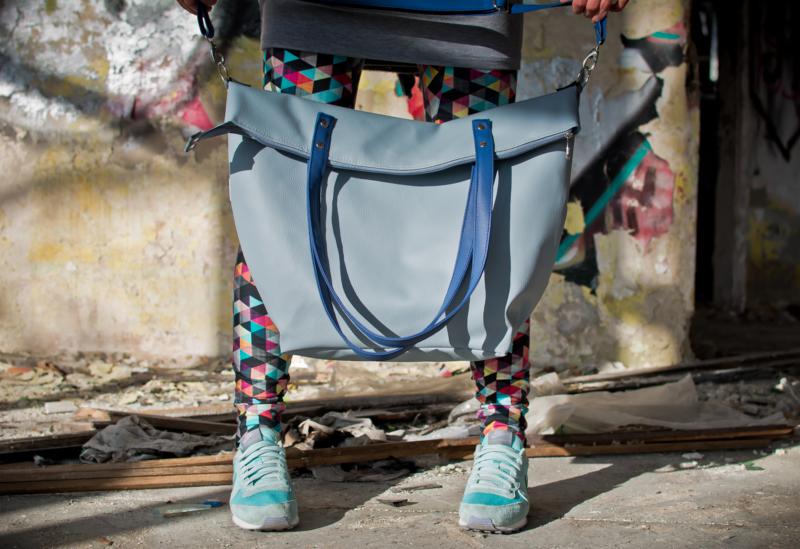 Jasnoniebieska torba shopper z granatowymi uchwytami, trzymana przez osobę w legginsach w geometryczne wzory i sportowych butach na tle ściany z graffiti.