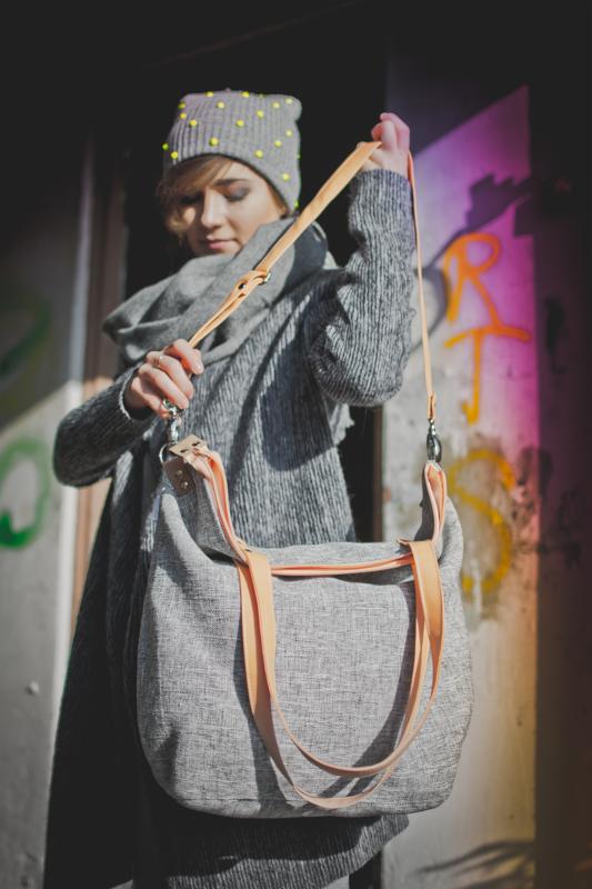 Szara torba z pomarańczowymi paskami, prezentowana przez kobietę w szarym stroju i czapce z żółtymi kropkami na tle ściany z graffiti. Ujęcie od przodu, skupione na detalu torby.