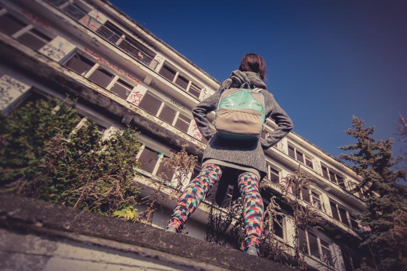 Kobieta z plecakiem stoi przed opuszczonym budynkiem z graffiti. Ubrana w wzorzyste legginsy i szary płaszcz. Widok z dołu, błękitne niebo w tle.
