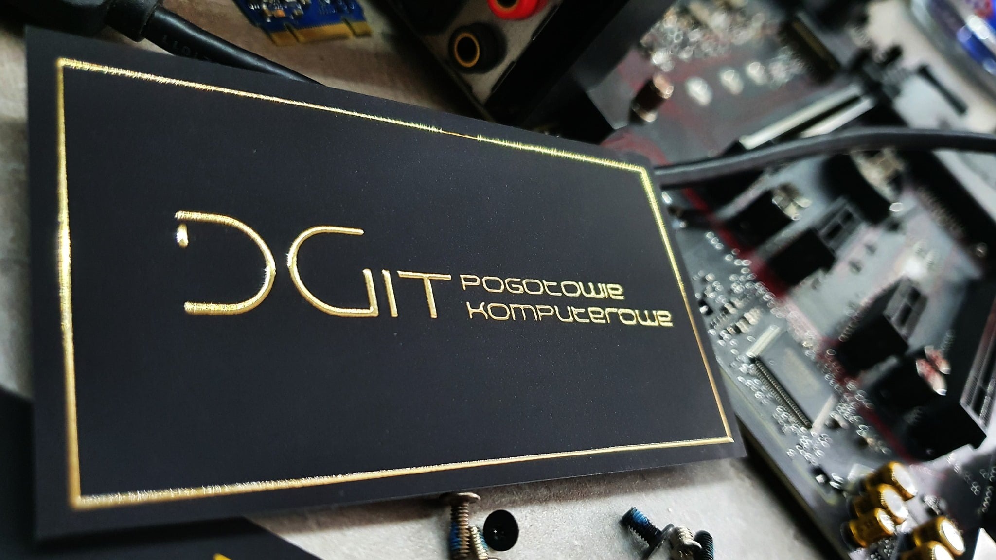Czarna wizytówka z napisem 'DGIT Pogotowie Komputerowe' w kolorze złotym, leżąca na tle rozmontowanej płyty głównej i śrubek.