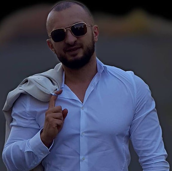 Portret mężczyzny w okularach przeciwsłonecznych, z marynarką przewieszoną przez ramię, na rozmytym tle.