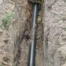 Czarna rura ułożona w wąskim wykopie w ziemi, widoczne połączenie rur z elementami montażowymi.