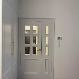 Białe drzwi wewnętrzne z szybkami, zamontowane w nowoczesnym wnętrzu z parkietem w jodełkę i białą szafą w zabudowie.