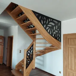 Drewniane schody ze spiralnym biegiem, ażurowe czarne balustrady z motywem liści, widok z dołu na jasne drewno i ciemną podłogę.