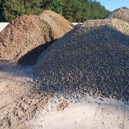 Usługi Sprzętowo Budowlane Ewa Źurawińska - Góra szarego gruzu na piaskowym podłożu, za nią wzniesienia z czerwonego i beżowego gruzu. W tle drzewa i błękitne niebo.