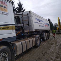 Usługi Sprzętowo Budowlane Ewa Źurawińska - Ciężarówka z naczepą Schmitz CargoBull na błotnistym terenie budowy, w tle koparki i pracownik w kamizelce odblaskowej. Widoczne reklamy usług transportowych i budowlanych.