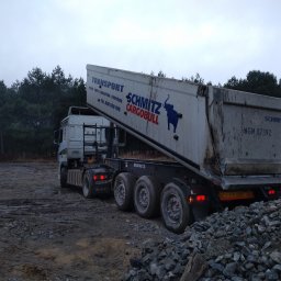 Usługi Sprzętowo Budowlane Ewa Źurawińska - Ciężarówka wywrotka Schmitz CargoBull z podniesioną skrzynią ładunkową, wysypująca gruz na placu budowy. W tle las iglasty i pochmurne niebo.