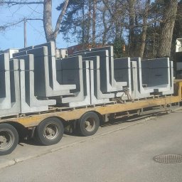 Usługi Sprzętowo Budowlane Ewa Źurawińska - Ciężarówka transportuje betonowe elementy L na lawecie, w tle drzewa i budynek. Widoczny dźwig i pracownik. Transport elementów prefabrykowanych.