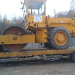Usługi Sprzętowo Budowlane Ewa Źurawińska - Żółty walec drogowy na niskopodwoziowej platformie transportowej. Widoczne opony, kabina i fragment lasu w tle. Maszyna gotowa do transportu na plac budowy.