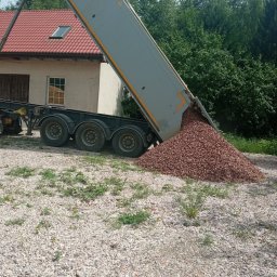 Usługi Sprzętowo Budowlane Ewa Źurawińska - Ciężarówka z uniesioną naczepą wysypuje brązowy gruz na jasne kamienie. W tle budynek z czerwonym dachem i zieleń drzew.