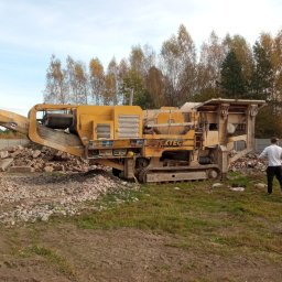 Usługi Sprzętowo Budowlane Ewa Źurawińska - Żółty mobilny kruszarka EXTEC na gąsienicach, obok sterta gruzu. W tle drzewa i betonowe ogrodzenie. Mężczyzna w białej koszulce stoi obok maszyny.